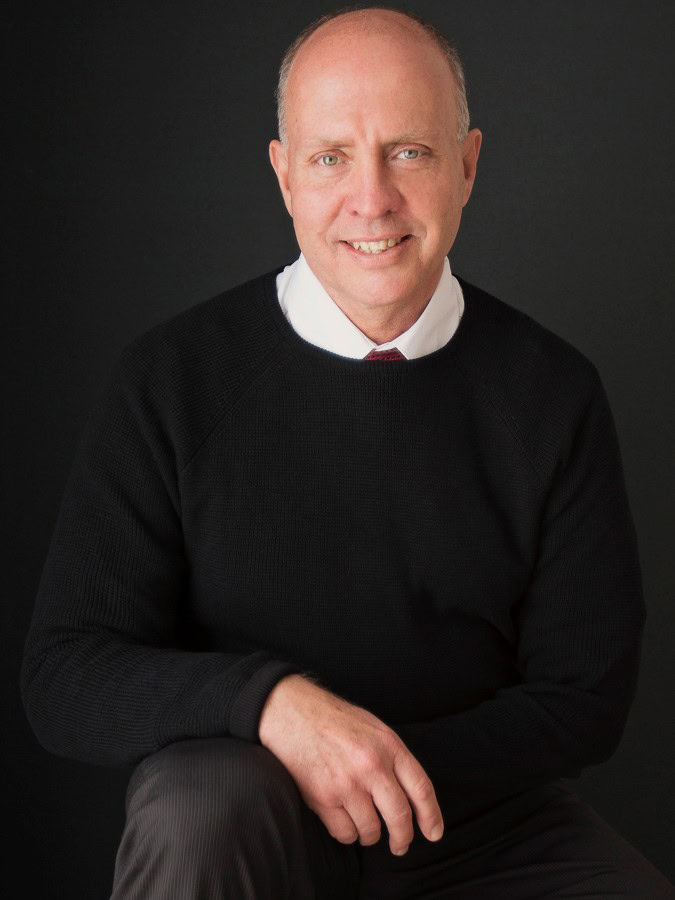 Portrait of a friendly middle-aged man in a black sweater and white dress shirt smiling against a black background.